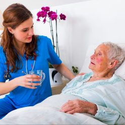 older woman reclining in bed with young woman in scrubs offering her a drink